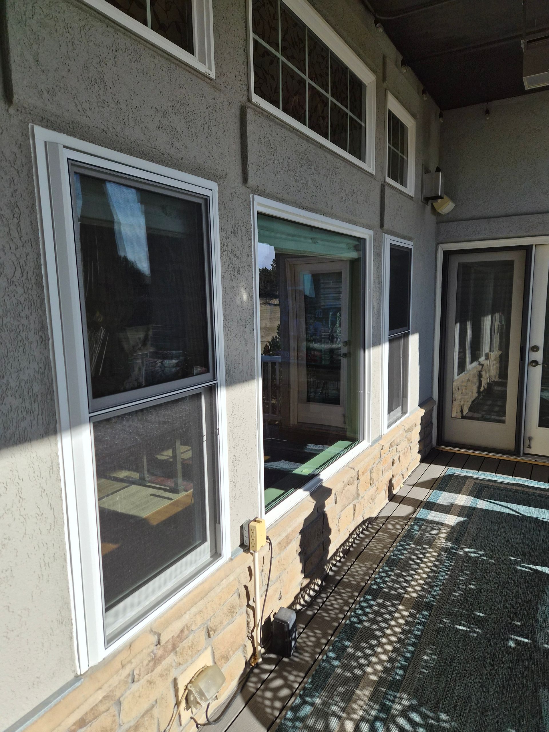 An outdoor patio area featuring a beige wall with multiple windows, a stone-patterned base, and a white screen door.
