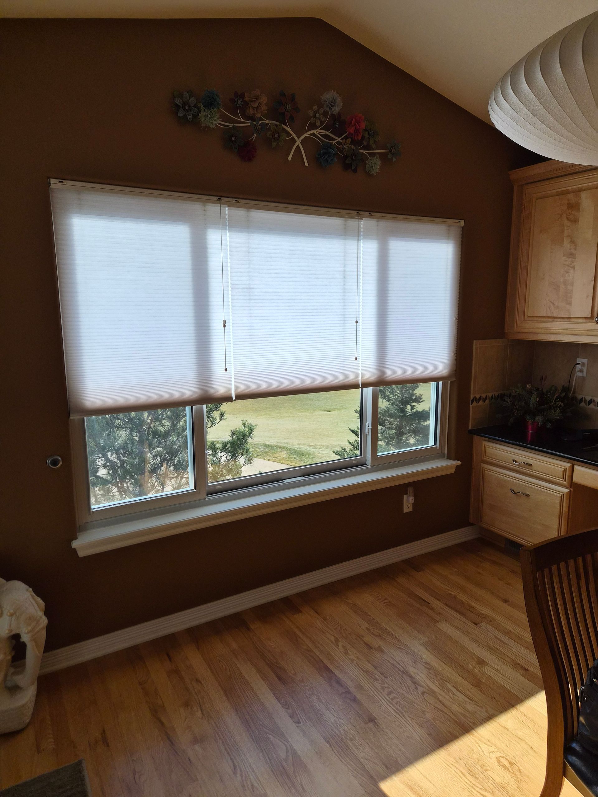 A wide window with white pleated blinds, overlooking trees, set in a brown-walled kitchen with wood cabinets and flooring.