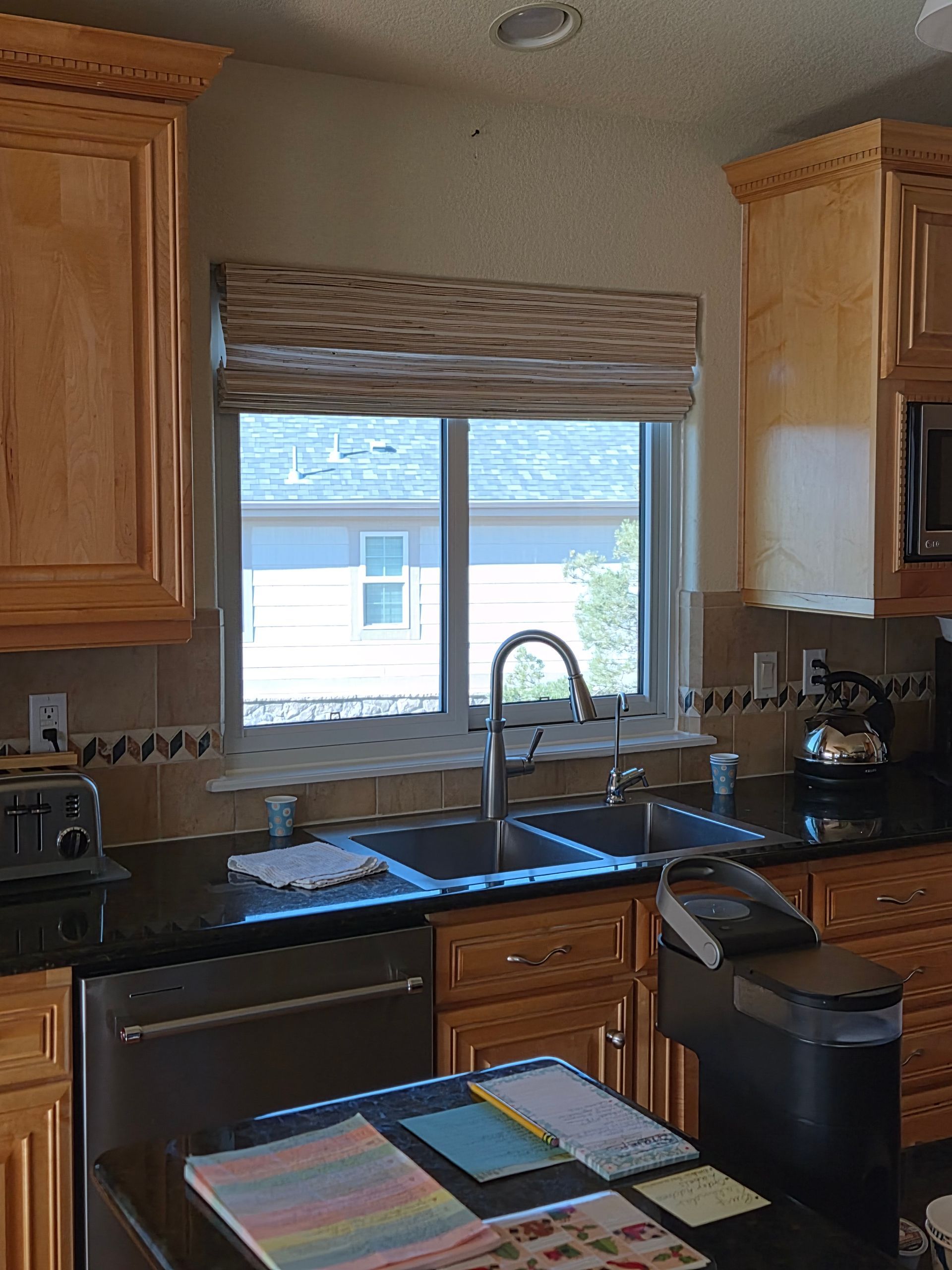 A kitchen sink area with light wood cabinets, dark countertops, a window with a woven shade, and a coffee machine.