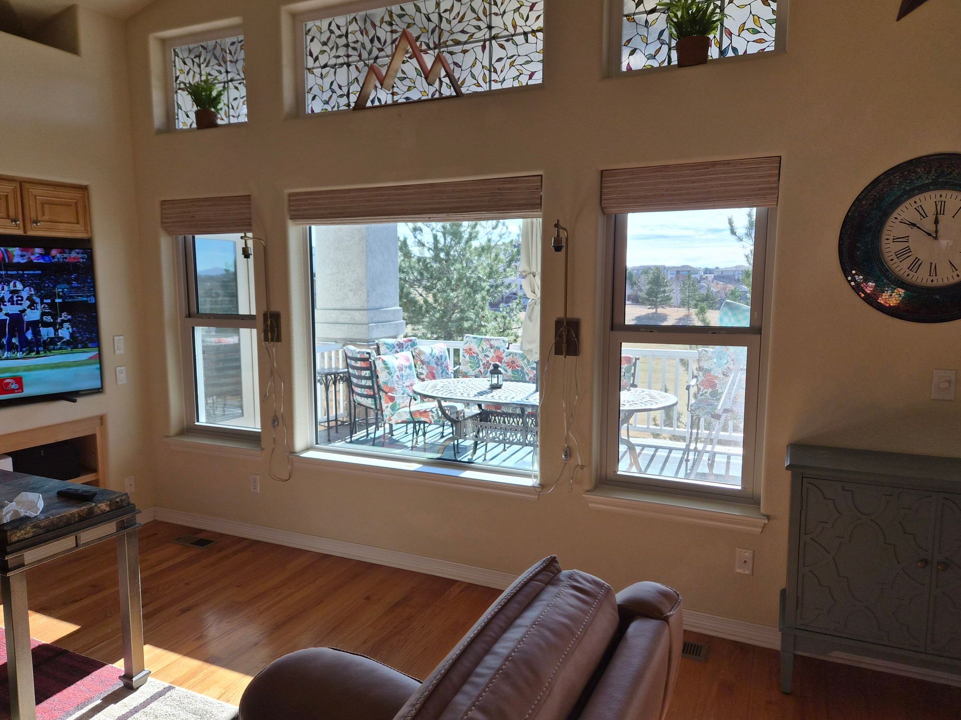 A living room with light walls, wood floors, large windows with woven shades, transom windows, a wall clock, and a TV.