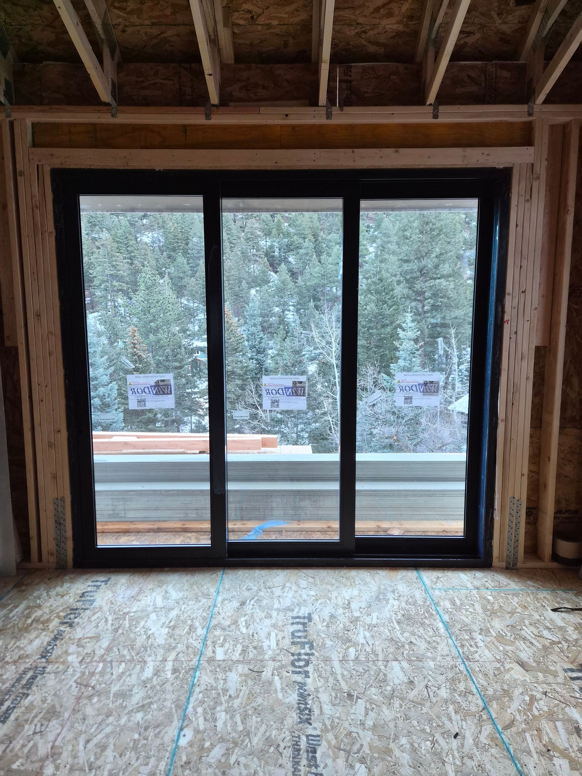 A large three-panel black sliding glass door installed in an unfinished room with wooden framing and OSB subflooring.