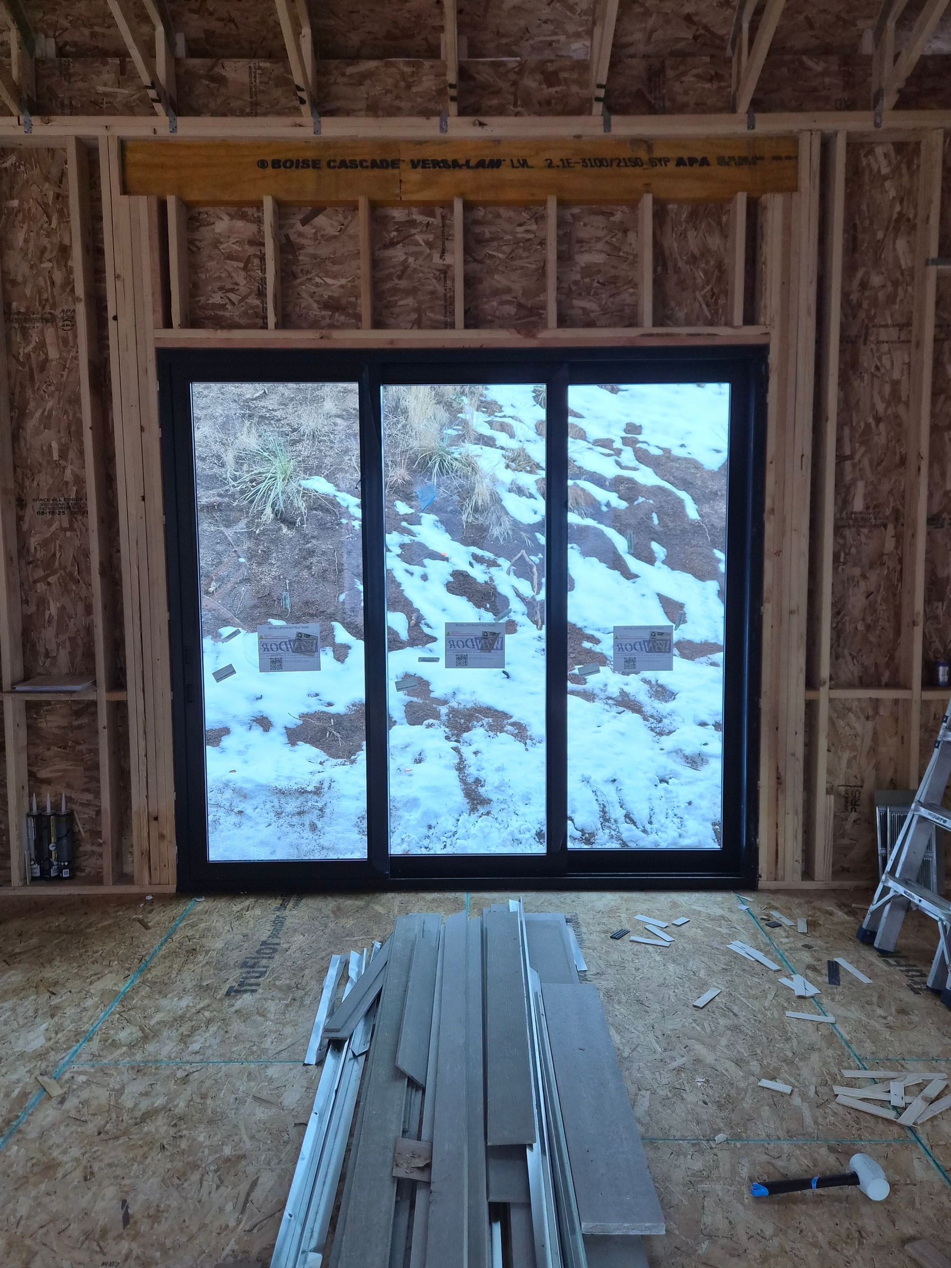 Black framed sliding glass door installed in a wooden, unfinished construction room looking out onto a snowy hill.