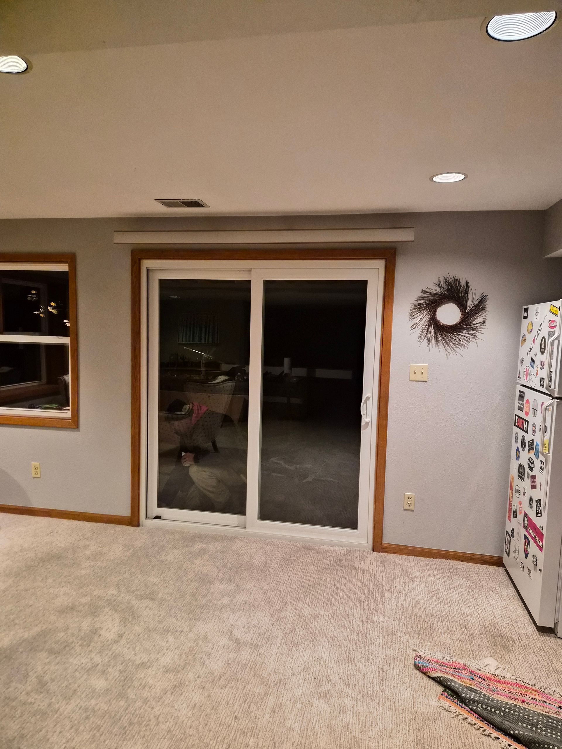 A room with gray walls, carpet, a sliding glass door with wood trim, a refrigerator to the right, and a wall wreath.