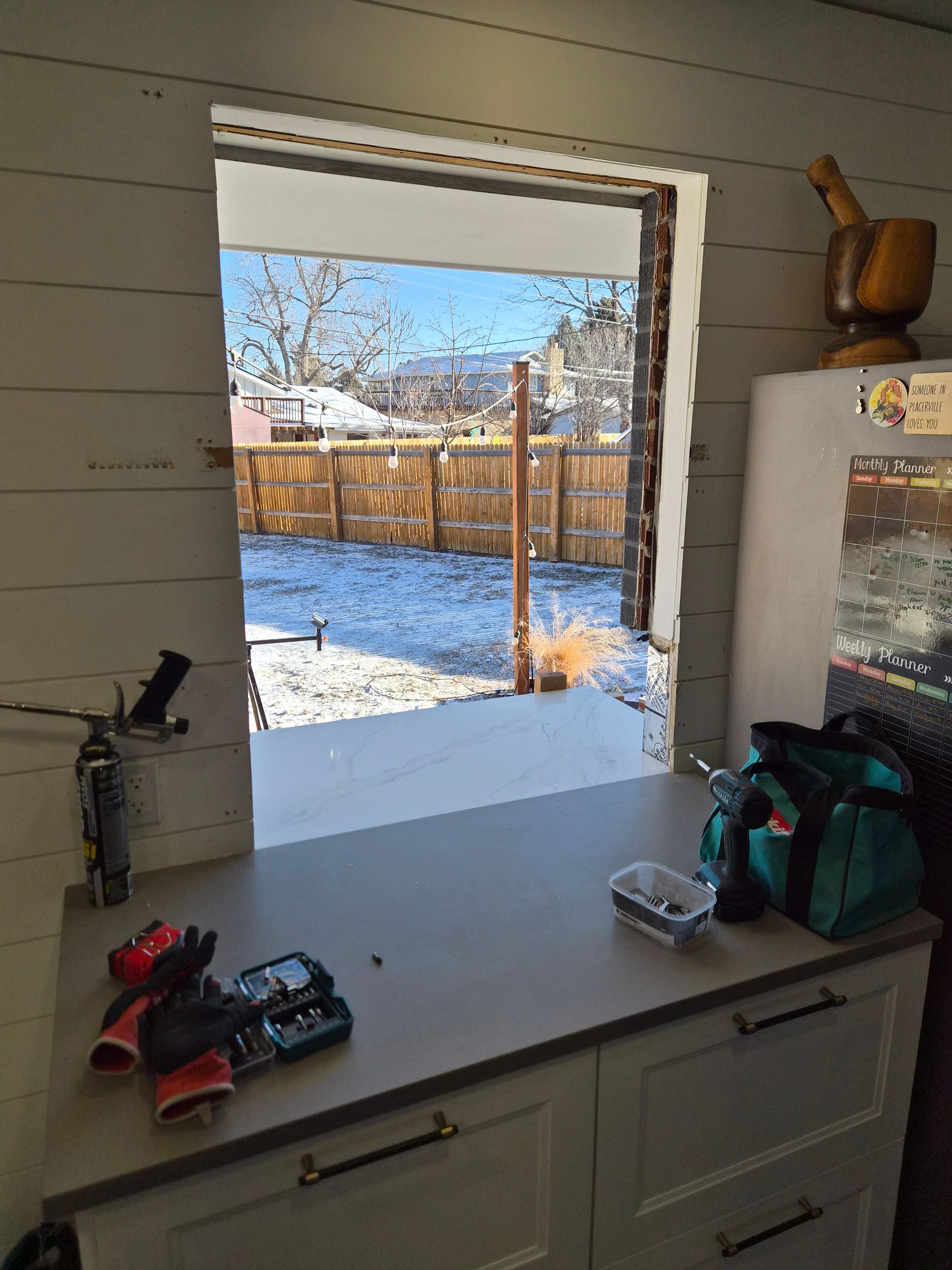 A rectangular opening in an interior wall reveals a snowy backyard with a wooden fence through a white-walled room.
