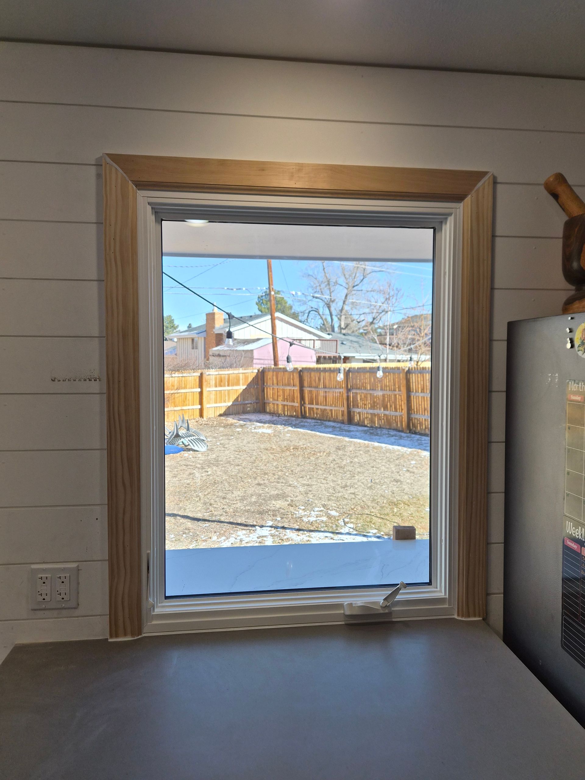 A rectangular window with light wood trim set into a white shiplap wall, looking out onto a fenced-in backyard.