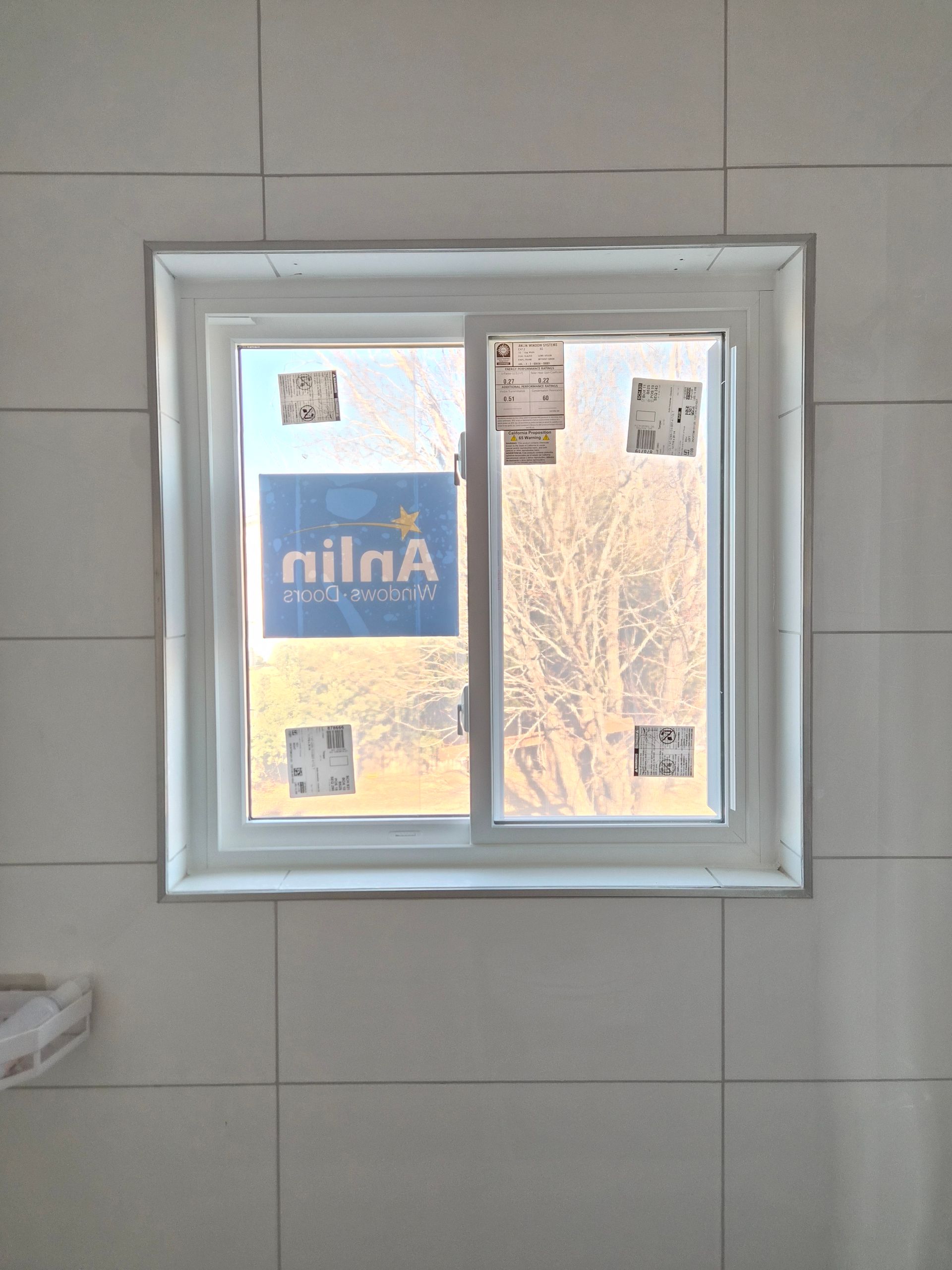 A white-framed sliding window installed in a tiled wall, with a partial product sticker visible on the glass.