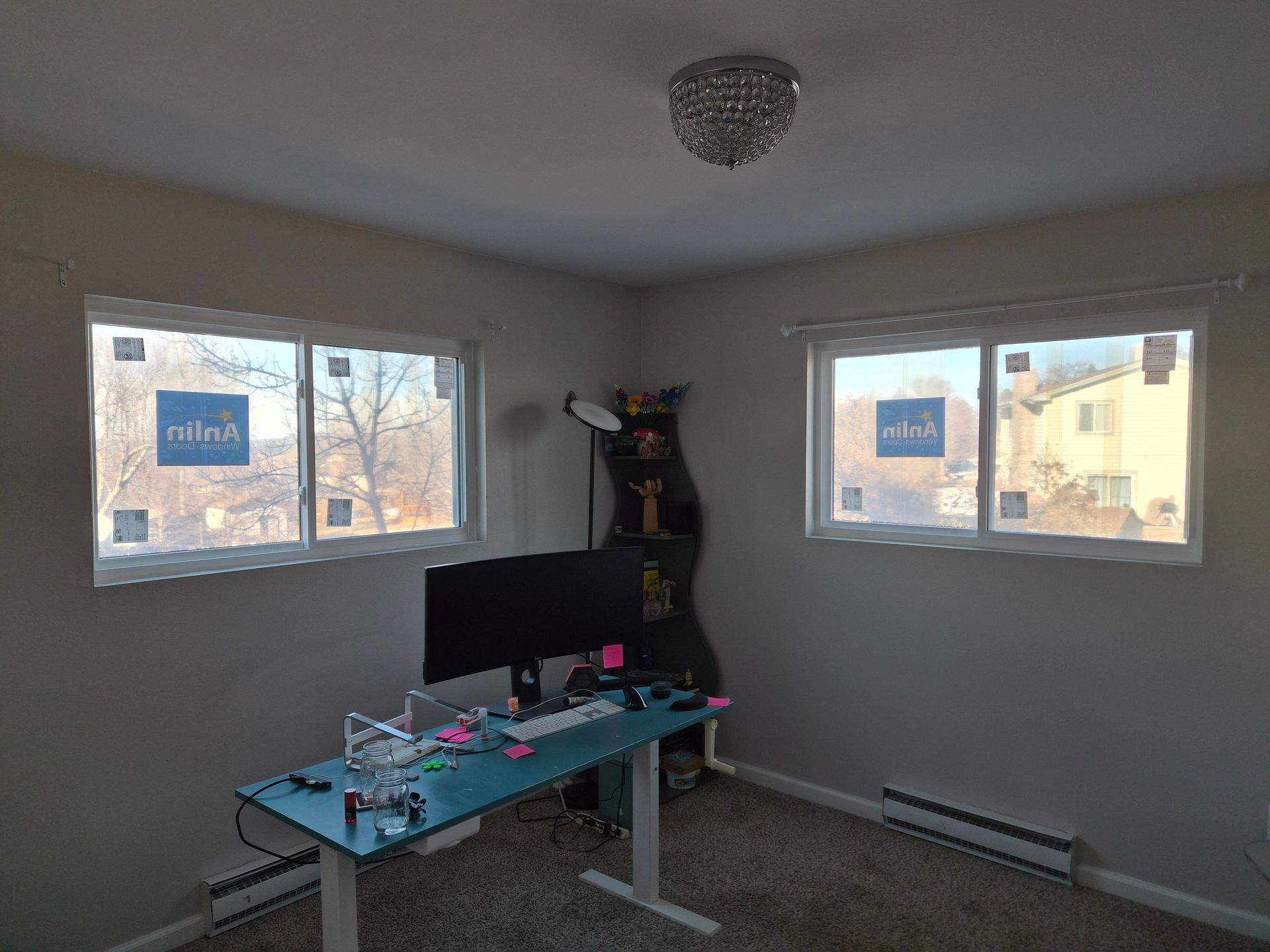 A corner home office with a desk, computer monitor, and two windows with blue brand stickers, centered on a gray wall.