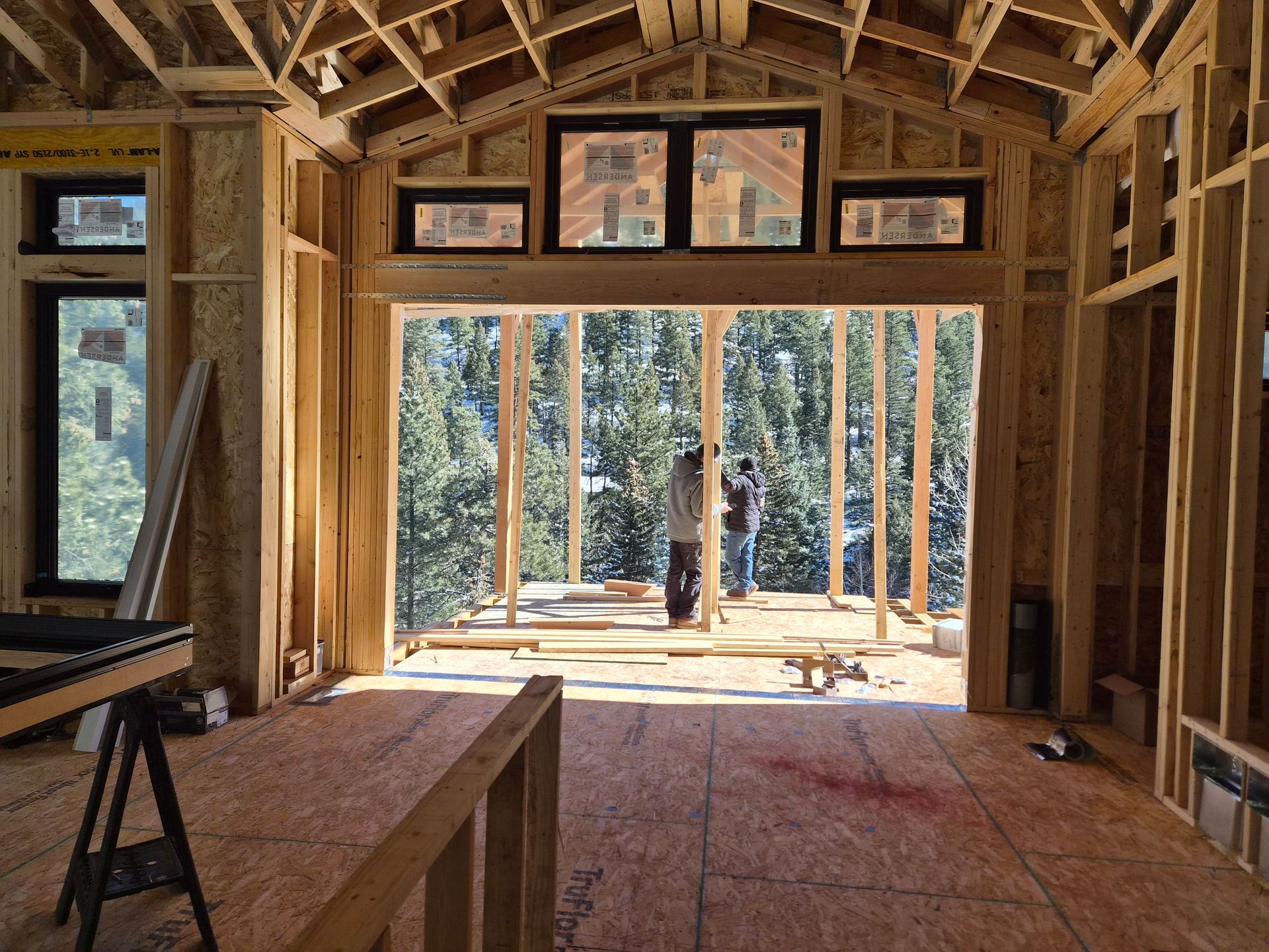 Interior view of a residential construction site looking out a large wall opening toward a forest.
