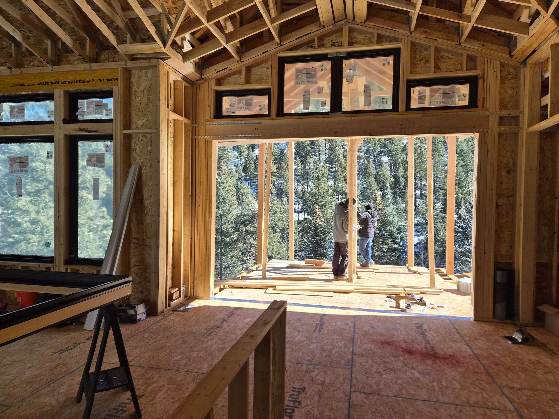 Construction site interior showing wooden framing, large window openings, and people working on a deck viewed from within.