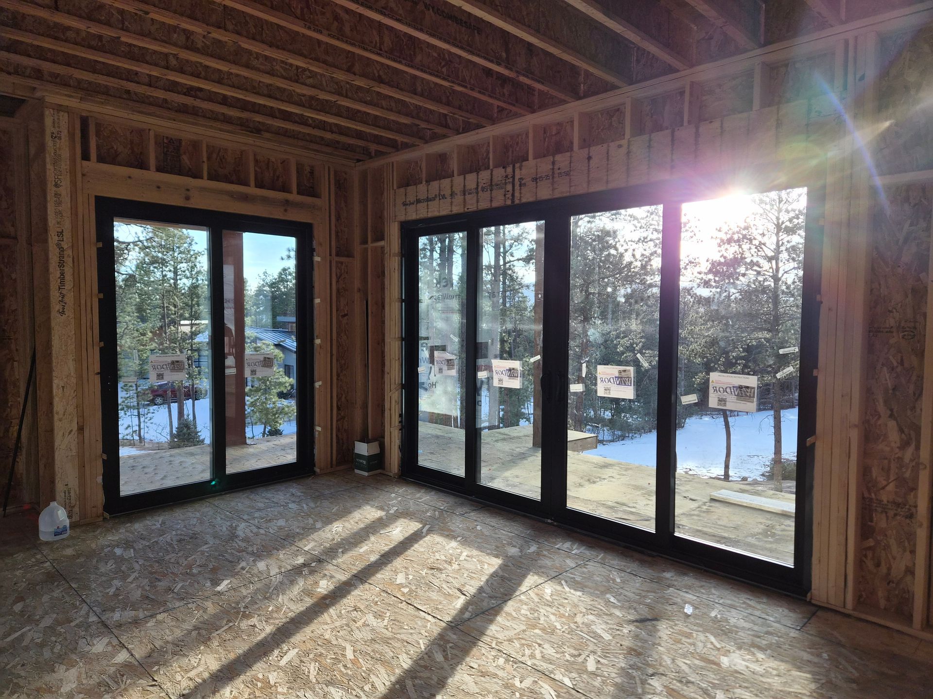 Unfinished room with black-framed glass sliding doors overlooking a snowy, treed landscape during construction.