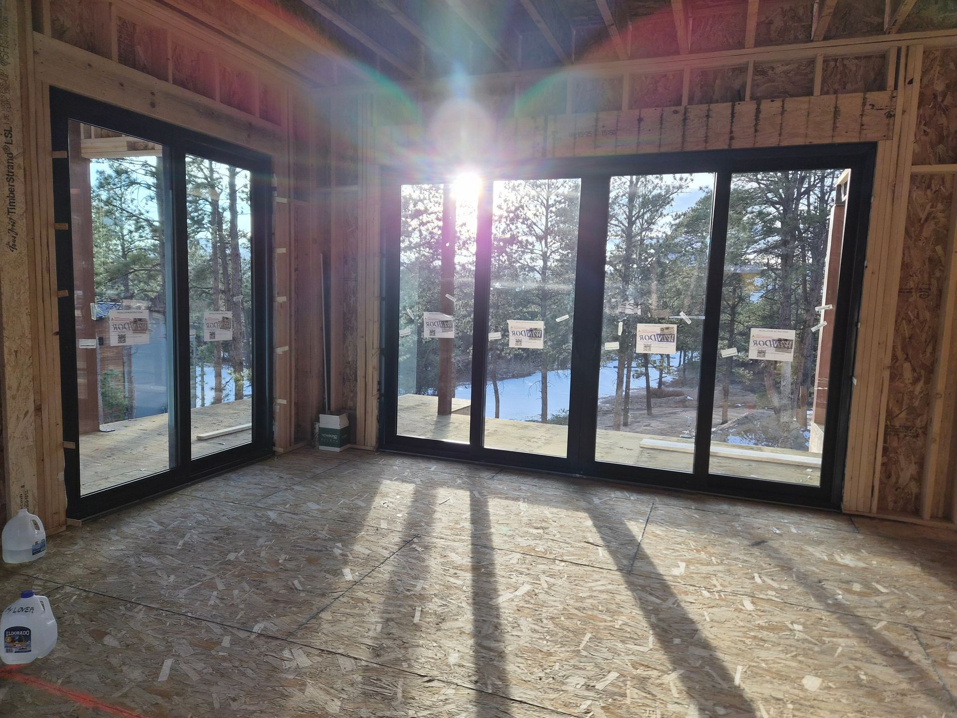 Interior view of a room under construction featuring large black-framed windows overlooking a snowy wooded area.