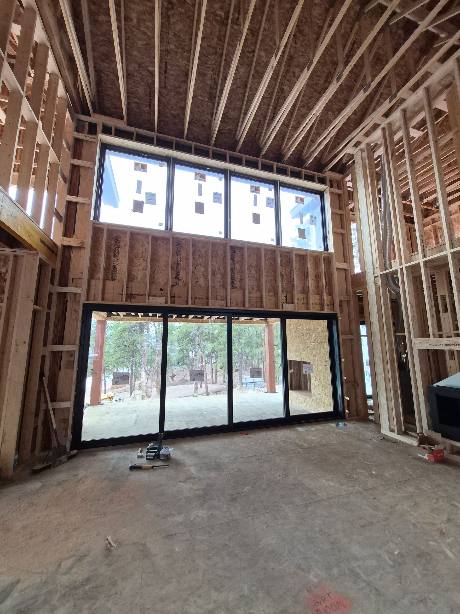 Interior view of a residential construction site featuring large glass sliding doors and upper windows framing a forest.