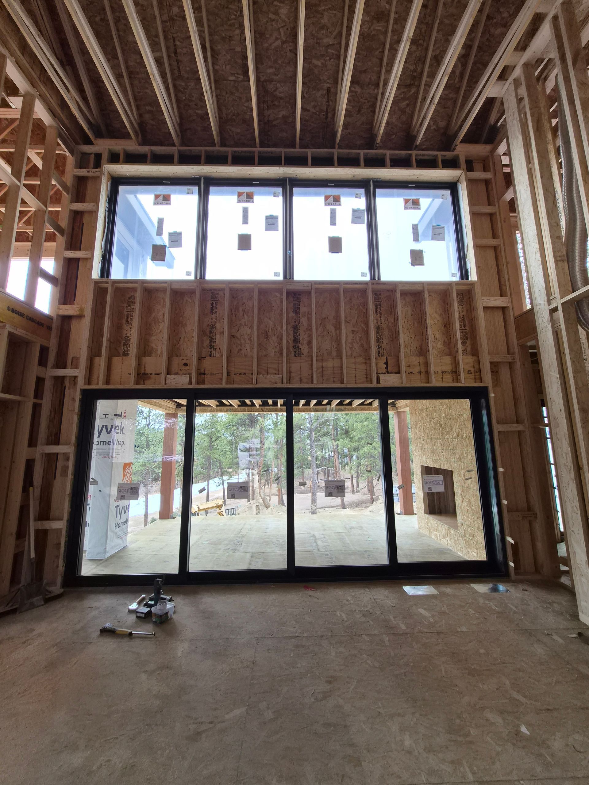 An interior view of a room under construction featuring large windows and sliding glass doors looking out to a forest.