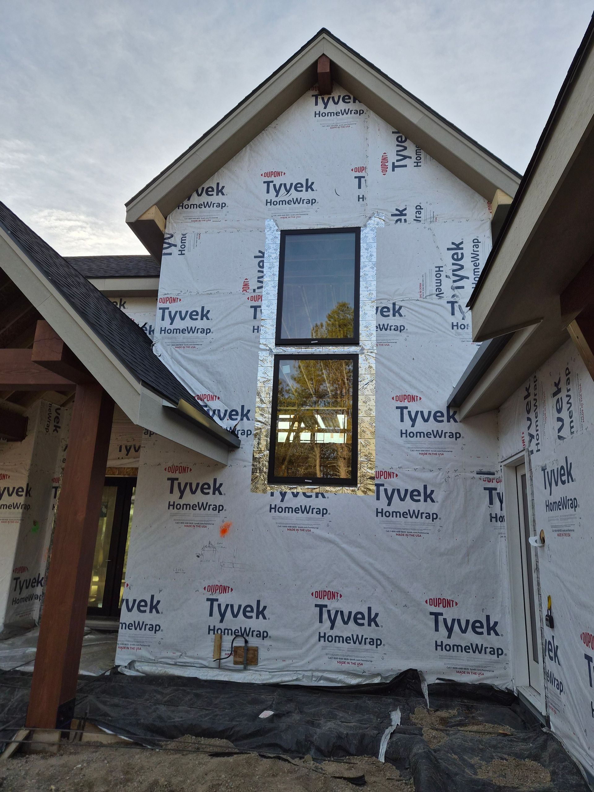 An unfinished house exterior with white Tyvek house wrap, wooden support beams, and a tall, two-pane black window frame.