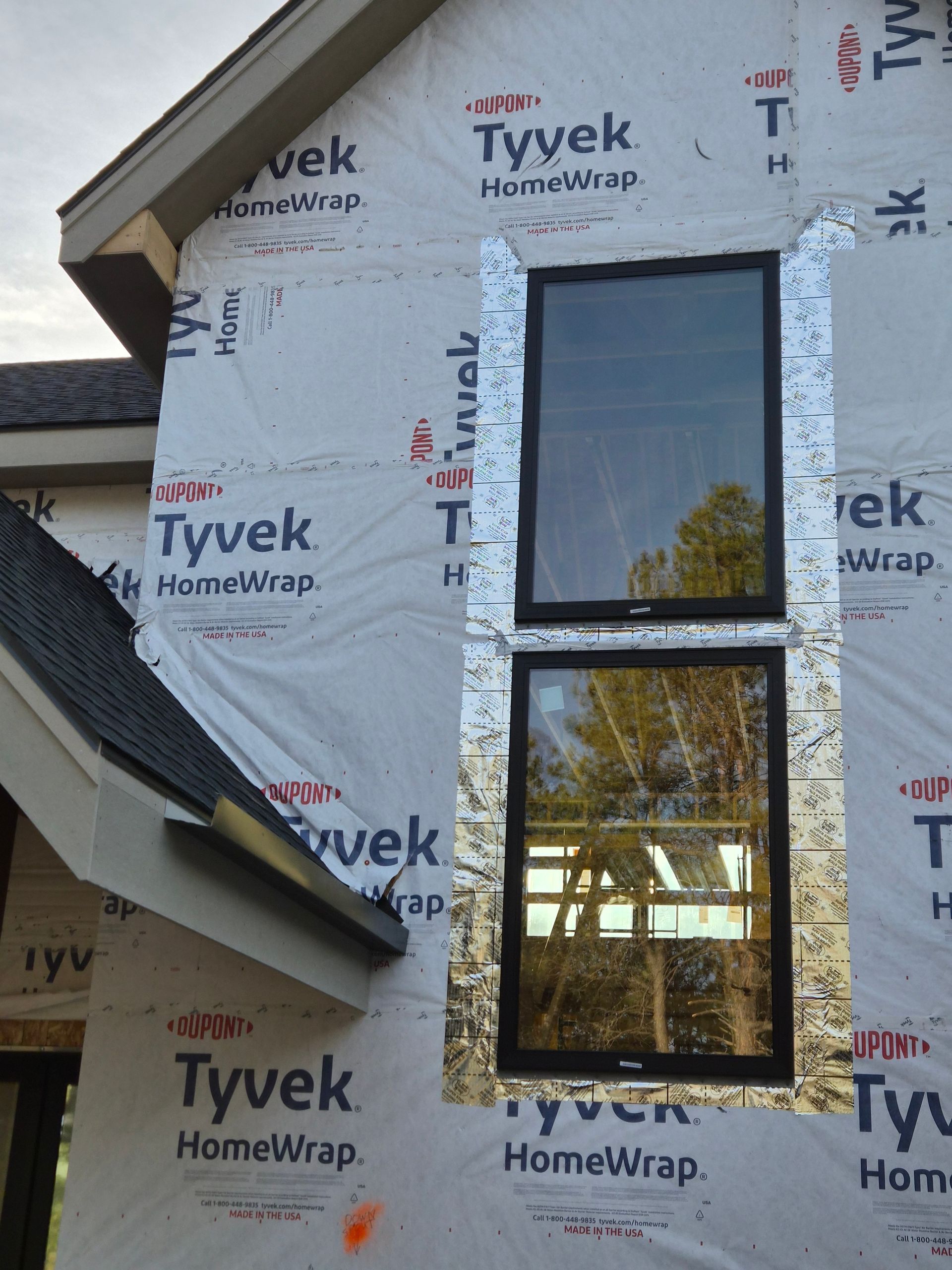 Two black-framed windows installed on a house exterior covered with Tyvek HomeWrap and metallic flashing tape.