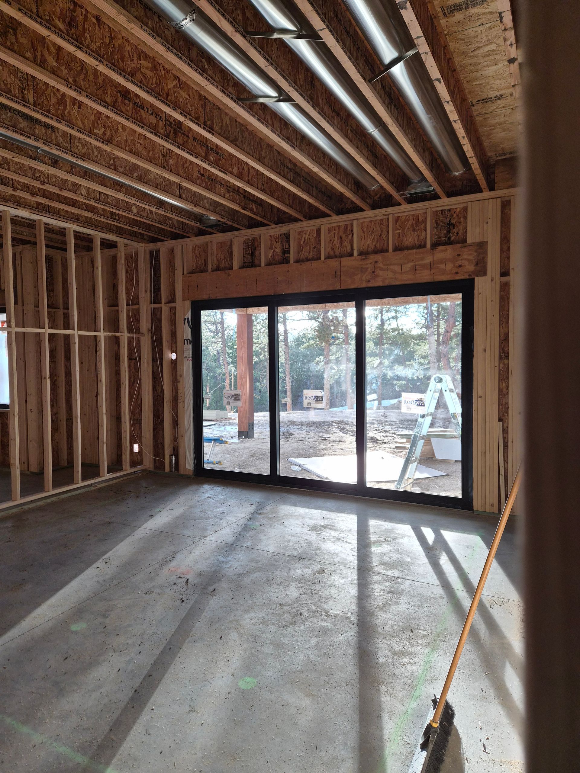 Interior of a room under construction with exposed wooden framing, concrete flooring, and a large black sliding door.