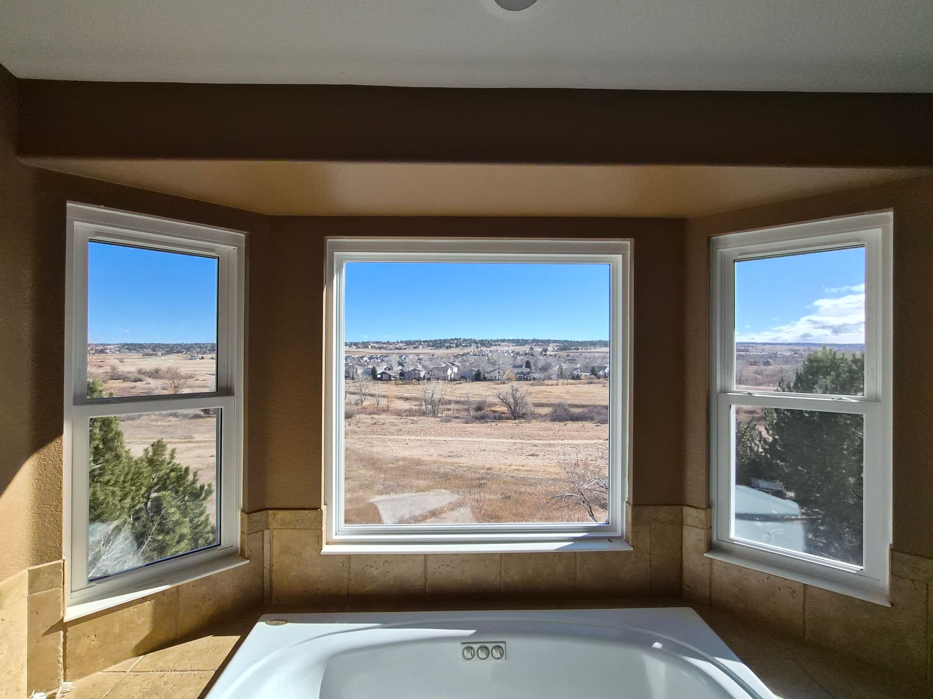 A bathtub sits in front of a bay window offering a view of a grassy field, a suburban neighborhood, and blue sky.