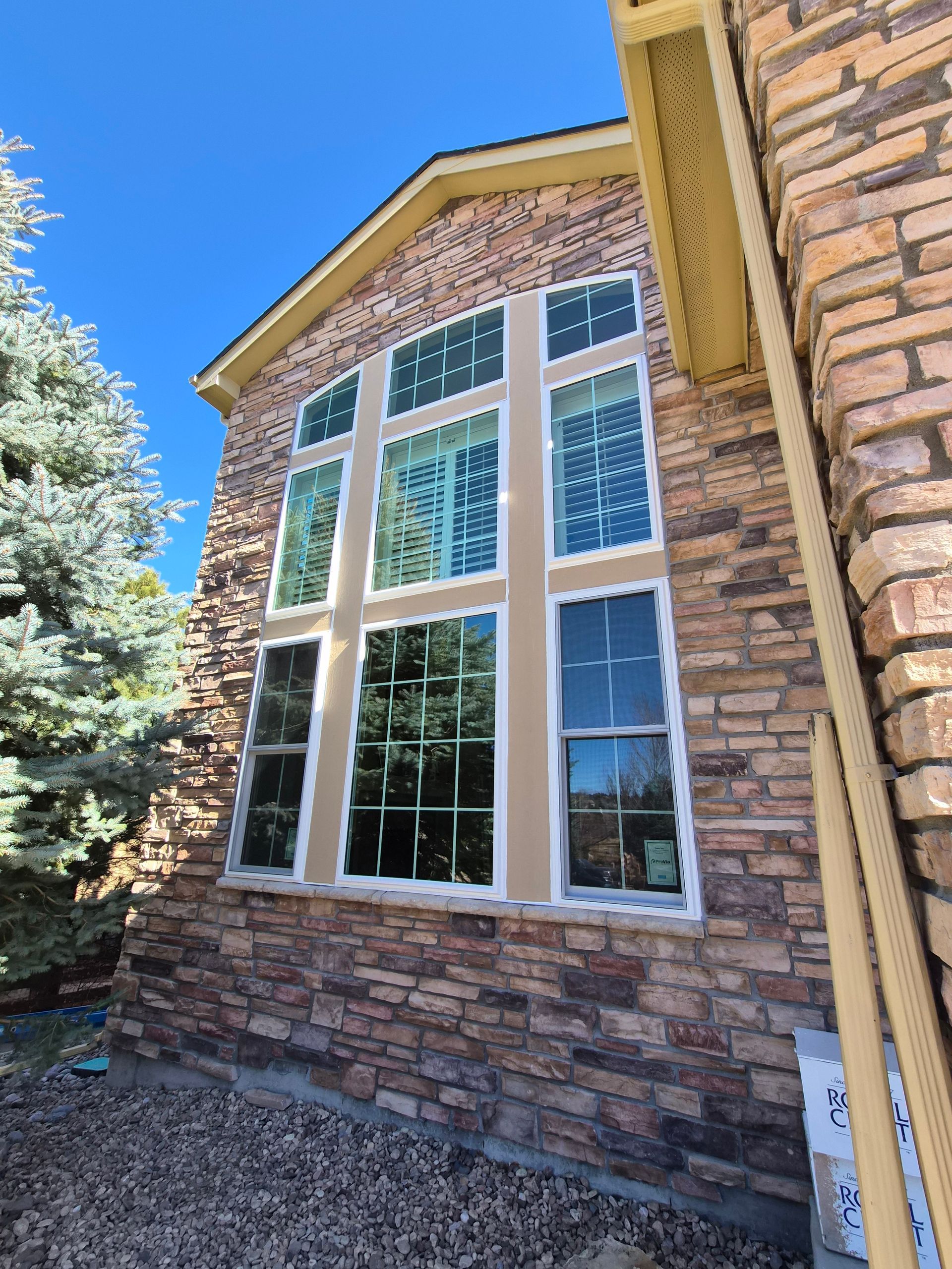 Large multi-pane window with a tan frame set into a rustic brown stone exterior wall of a house.