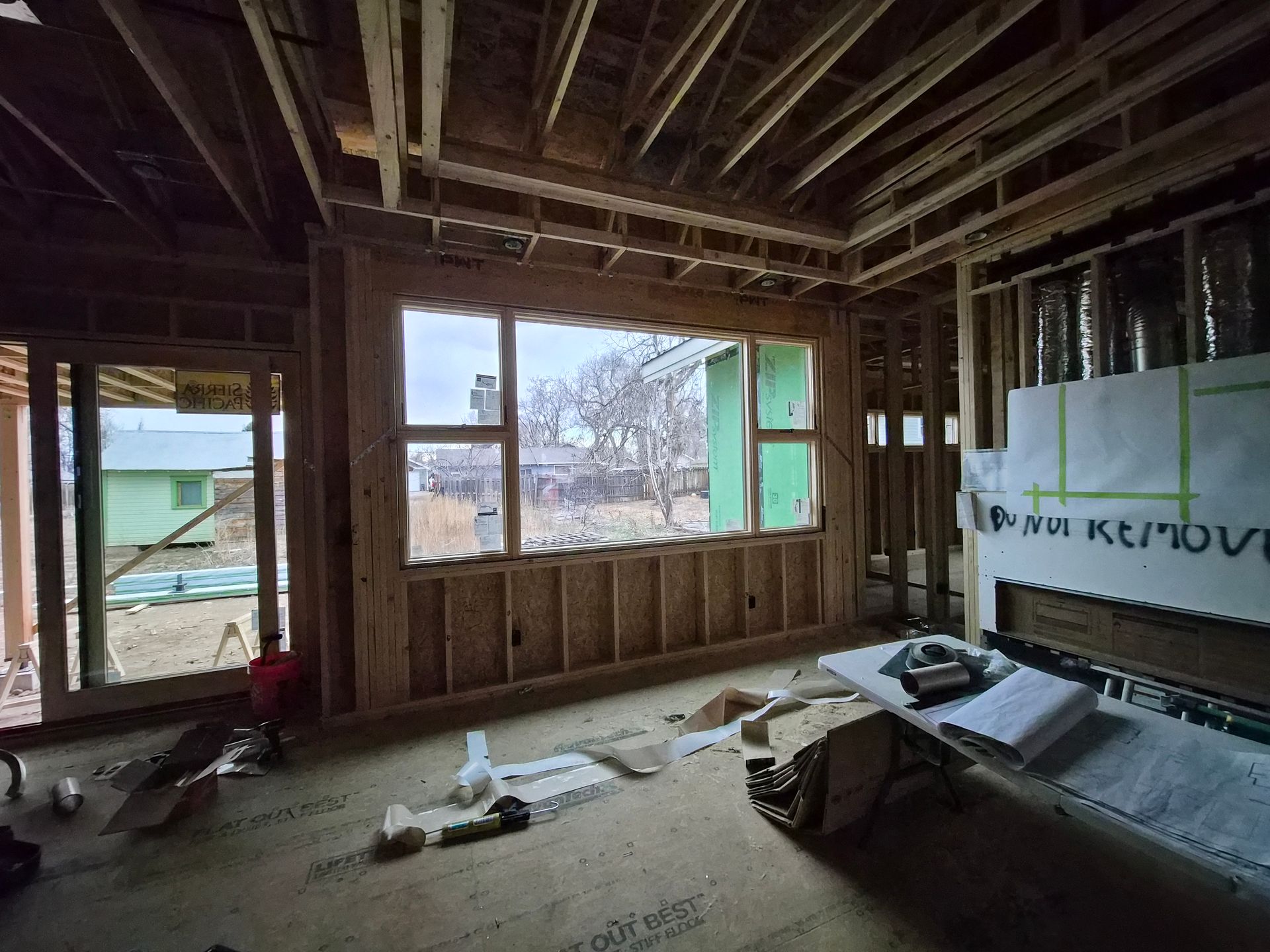 The interior of a house under construction with exposed wooden framing, a large window, and blueprints on a flat surface.