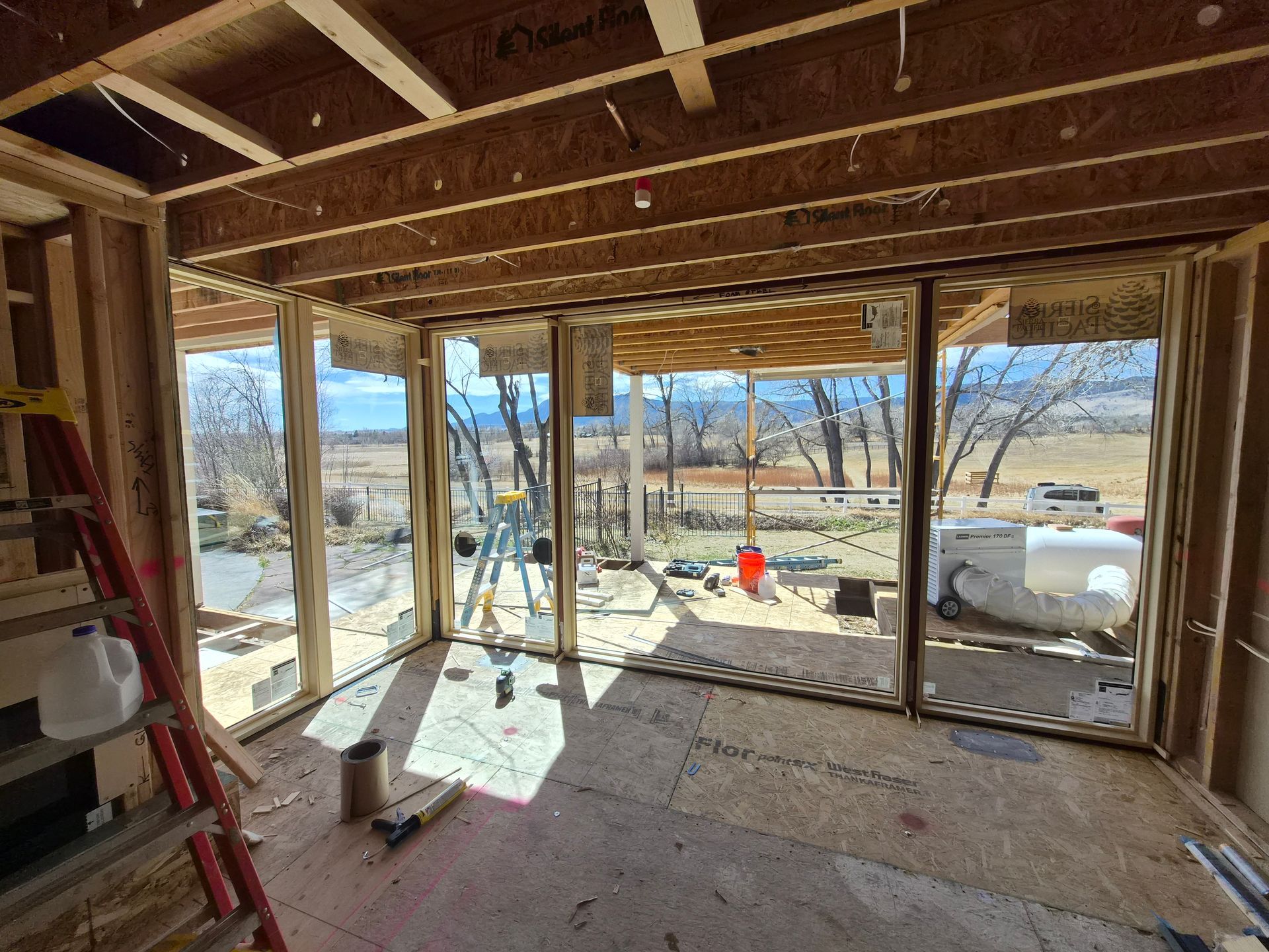 An unfinished room with exposed wooden framing and a large glass window overlooking a sunny, rural outdoor landscape.