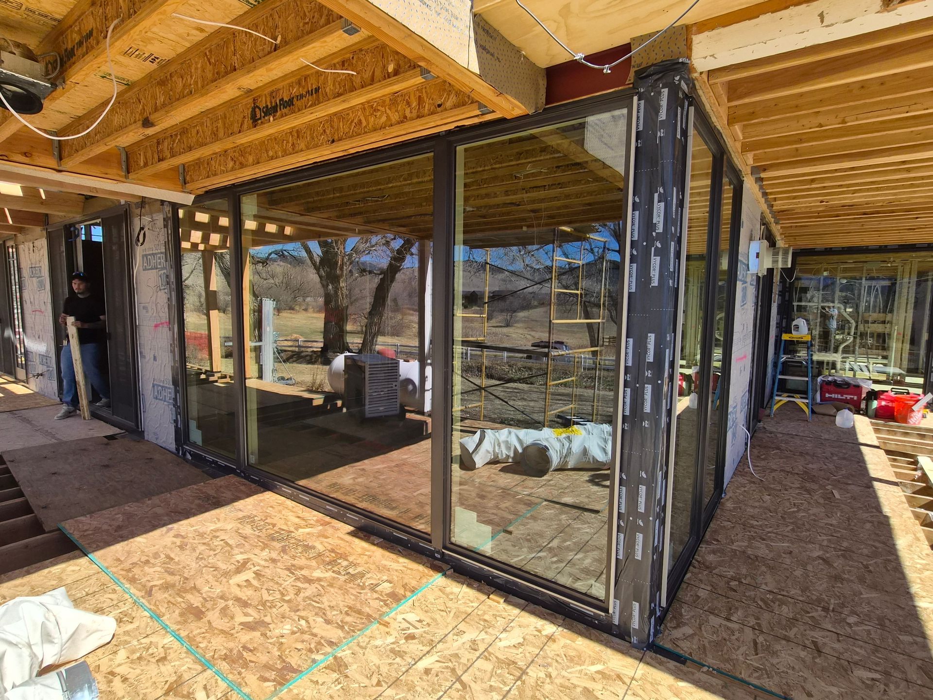 Construction site showing a house frame with large glass sliding doors installed and exposed plywood flooring.