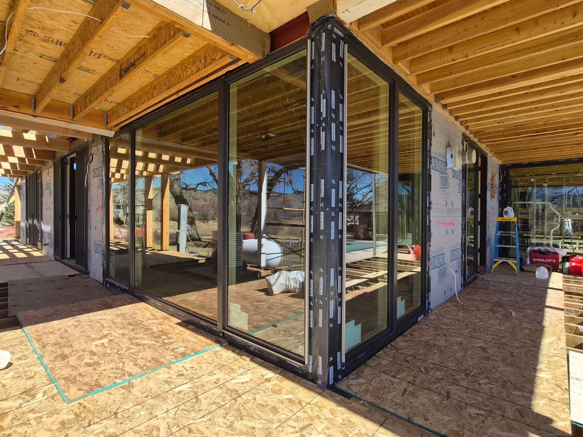 Modern construction site showing a corner glass window installation with exposed wooden framing and grey sheathing.