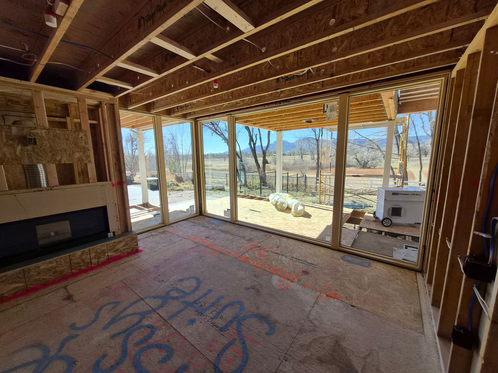 An interior view of a room under construction, with exposed wood framing, a fireplace, and large windows looking outside.