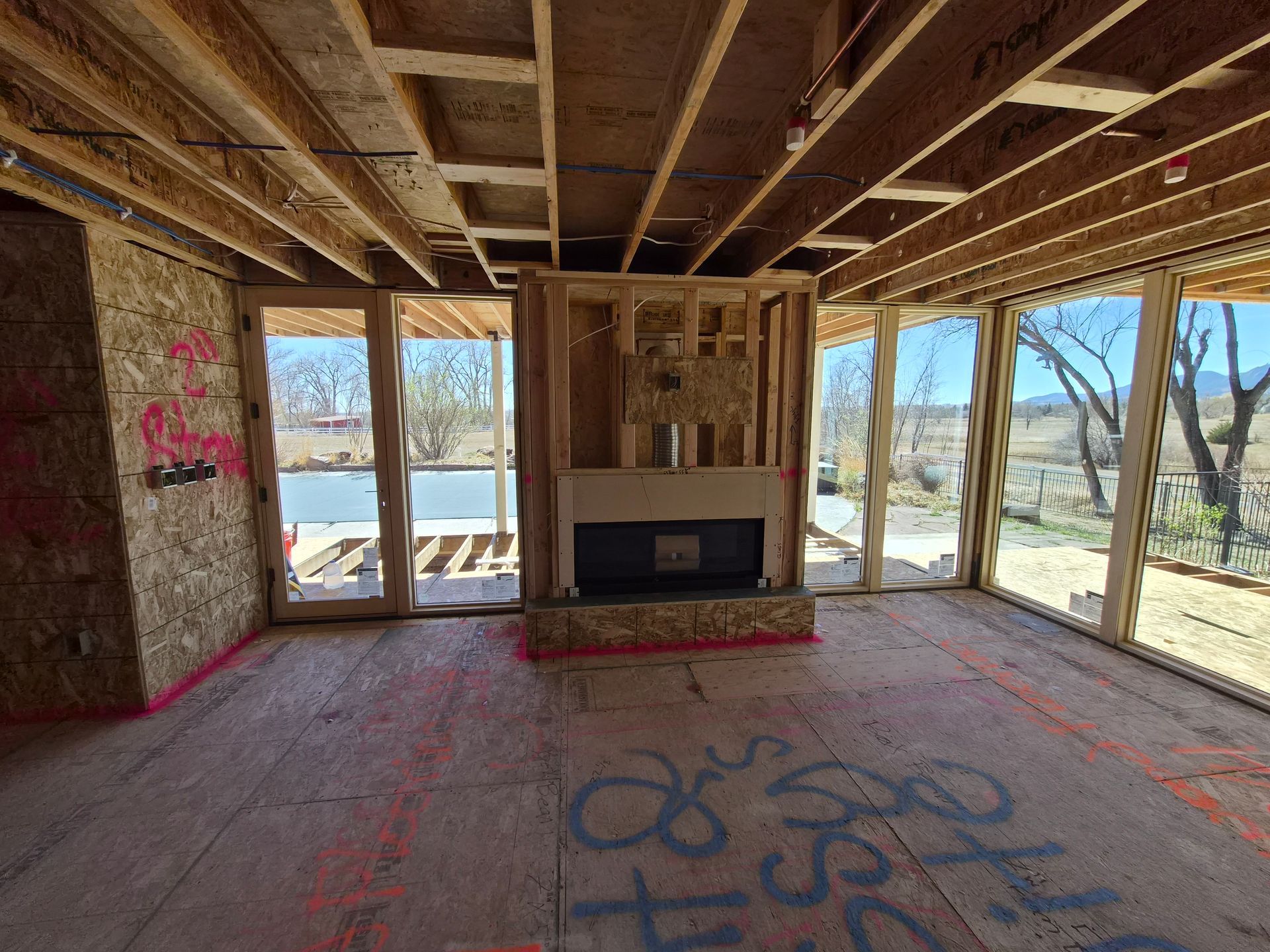 Interior construction of a room with a framed fireplace, bare wooden ceiling joists, and large windows overlooking a field.