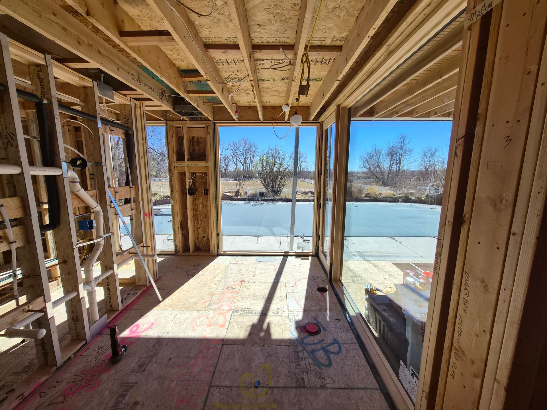 The interior of a house under construction with exposed wooden framing and large glass windows overlooking a field.