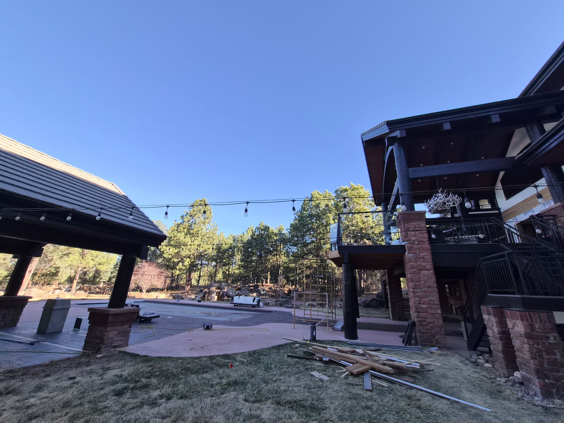 A sunny outdoor patio area between two rustic buildings, featuring brick pillars, covered porches, and pine trees.