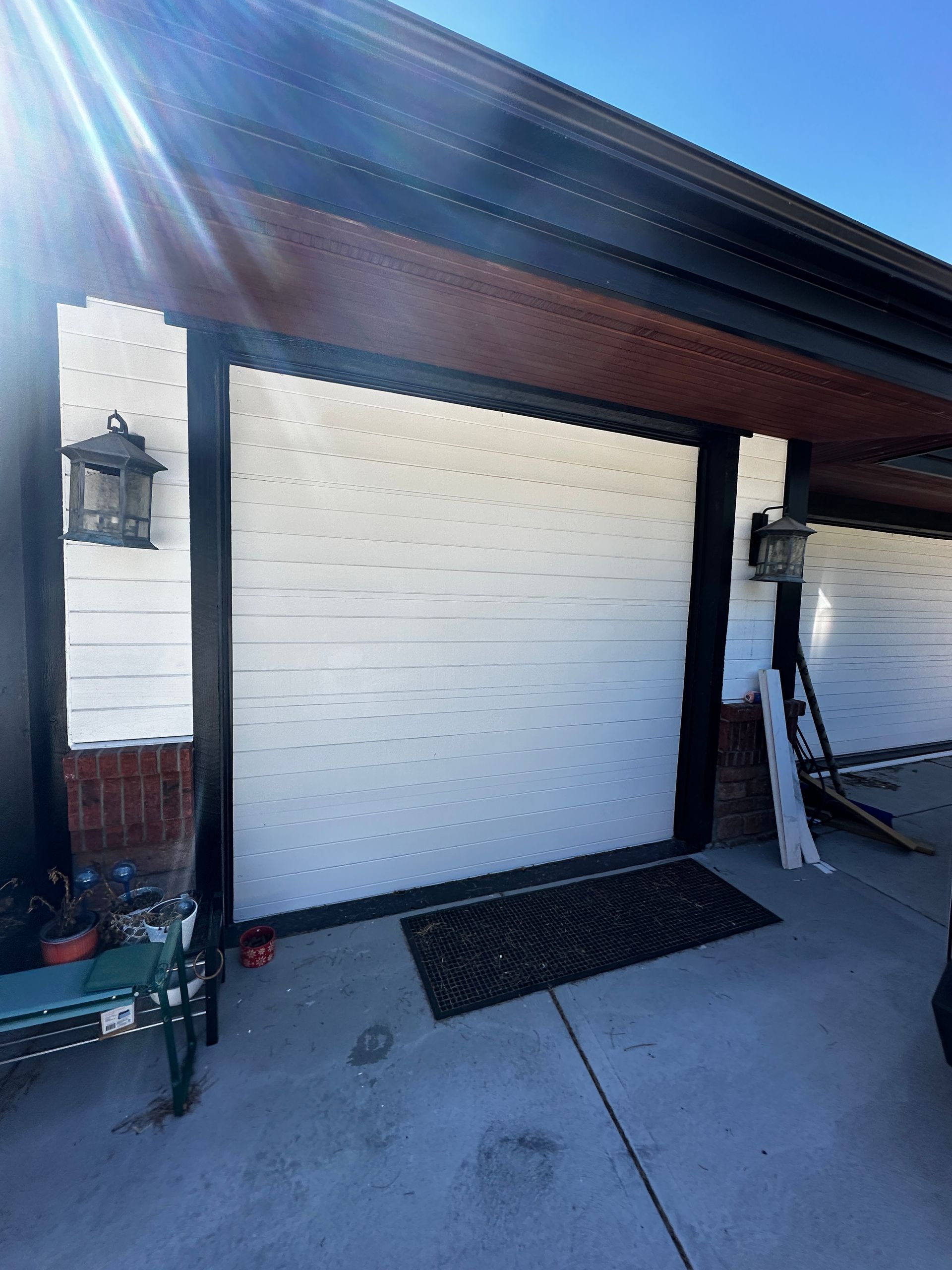 A white garage door with black trim and lanterns on either side, set in a white exterior wall with brick accents.
