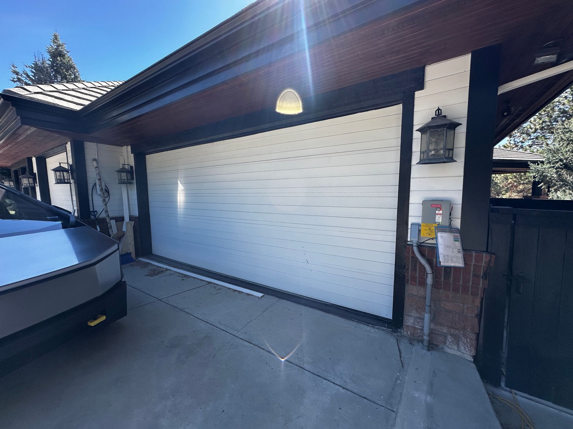 A white garage door and adjacent wall with an electrical meter, outdoor lantern, and a vehicle corner in a driveway.