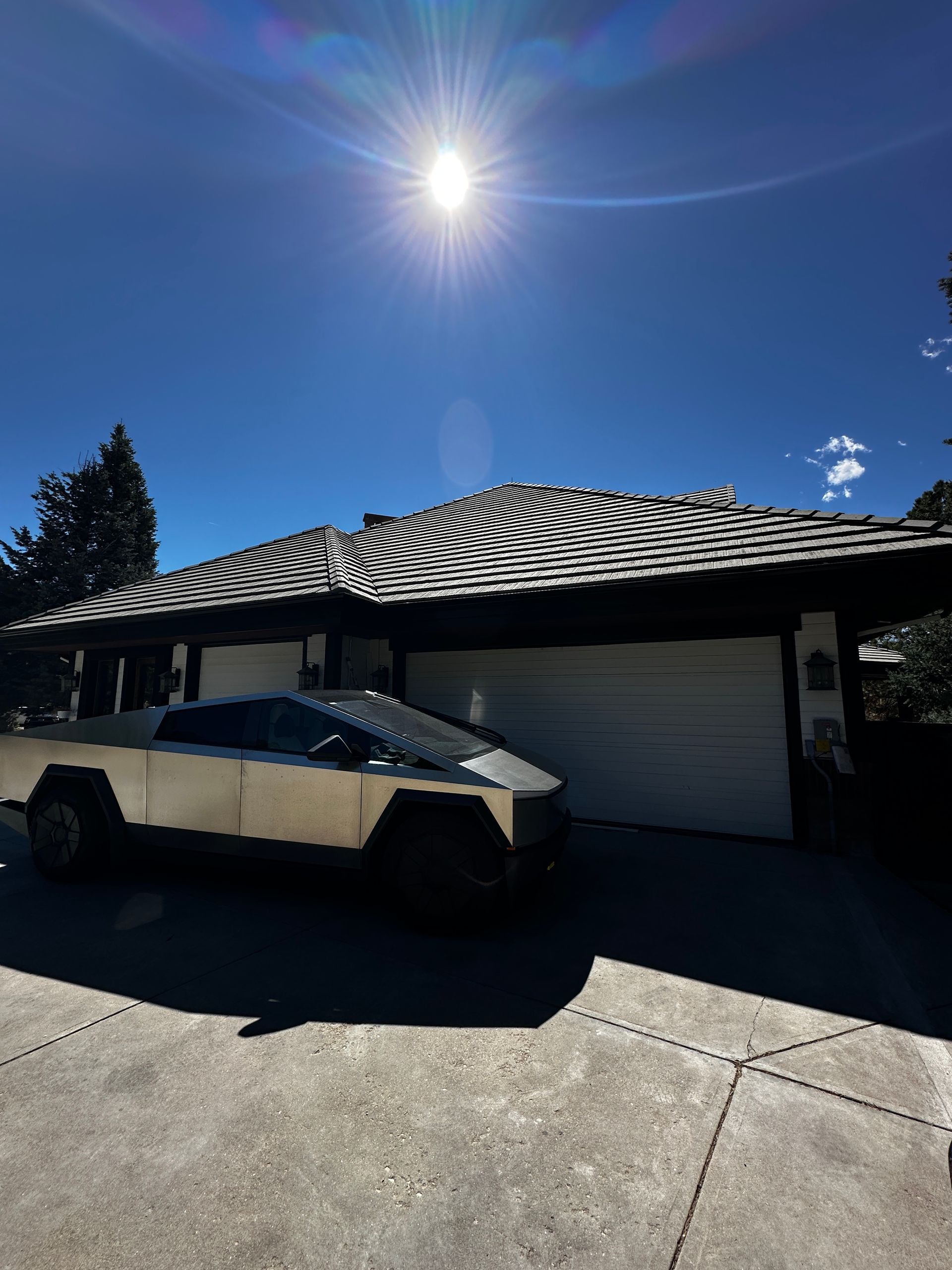 A silver Tesla Cybertruck parked on a driveway in front of a house with a patterned roof under a bright, sunny sky.