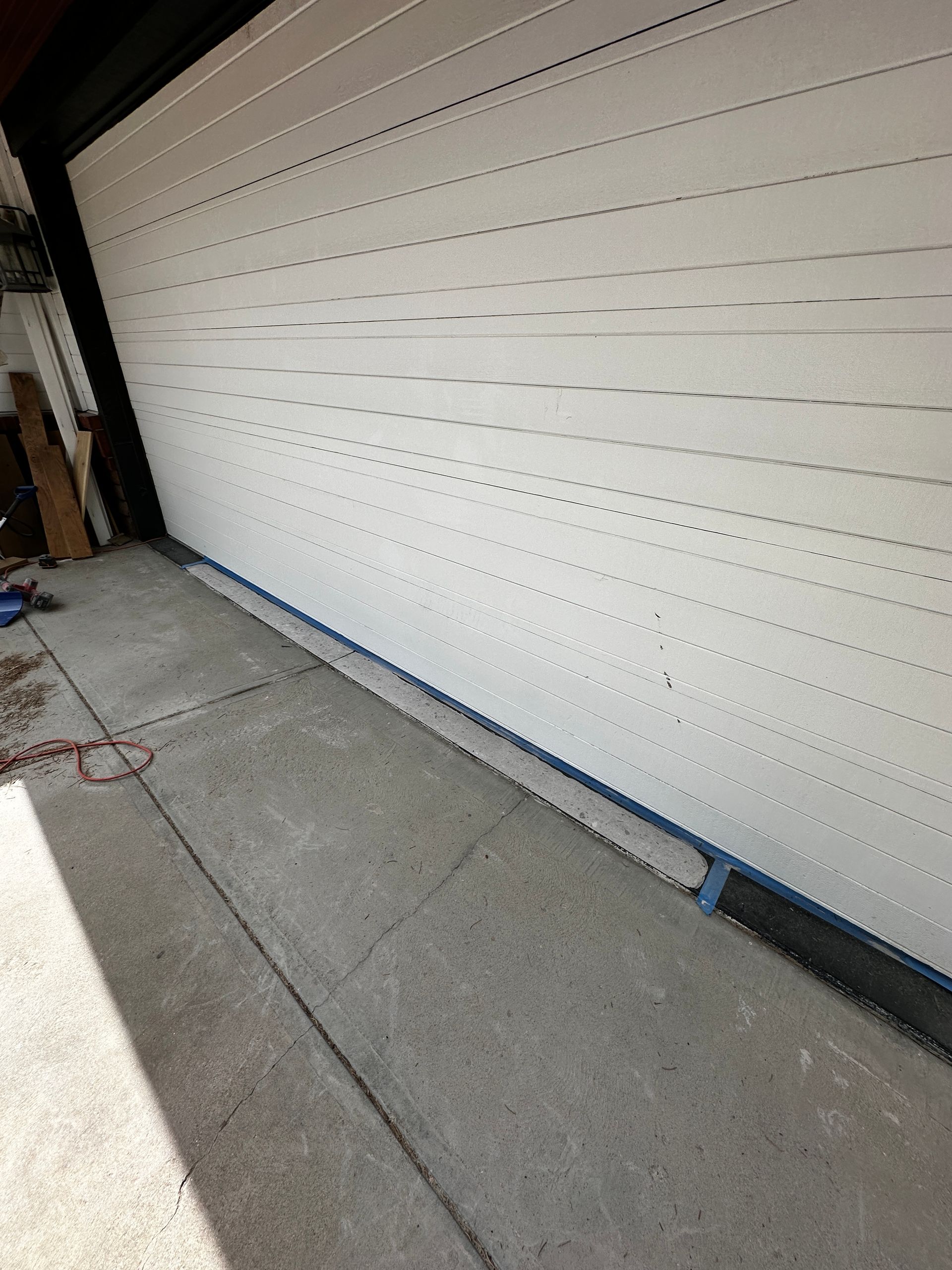 A beige garage door is closed against a concrete driveway, with a blue rubber weather seal strip along the bottom edge.