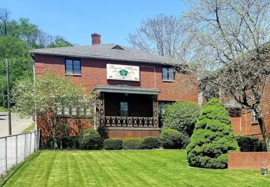 Brick building with a porch, green lawn, trees, and a sign that reads 