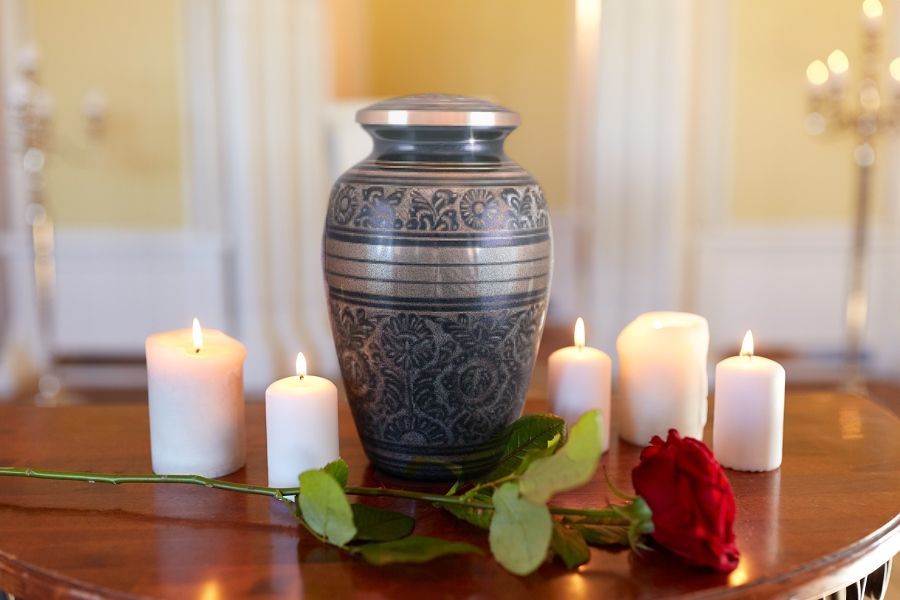 Urn with floral details, candles, and a rose on a table, likely in a memorial setting.
