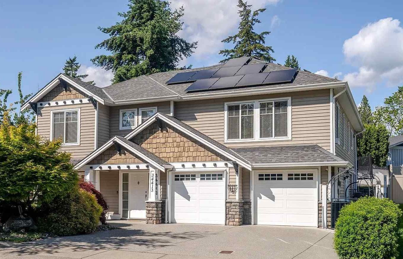 A two-story tan suburban house with solar panels on the roof and an attached two-car garage under a sunny blue sky.