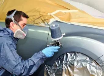 Operário com máscara de proteção respiratória pintando a carroceria de um carro em uma cabine de pintura.