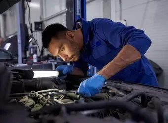 Mecânico de luvas azuis inspecionando o motor de um carro sob o capô com uma lanterna.