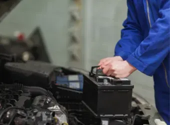 Pessoa com jaqueta de trabalho azul testando a bateria de um carro no compartimento do motor.