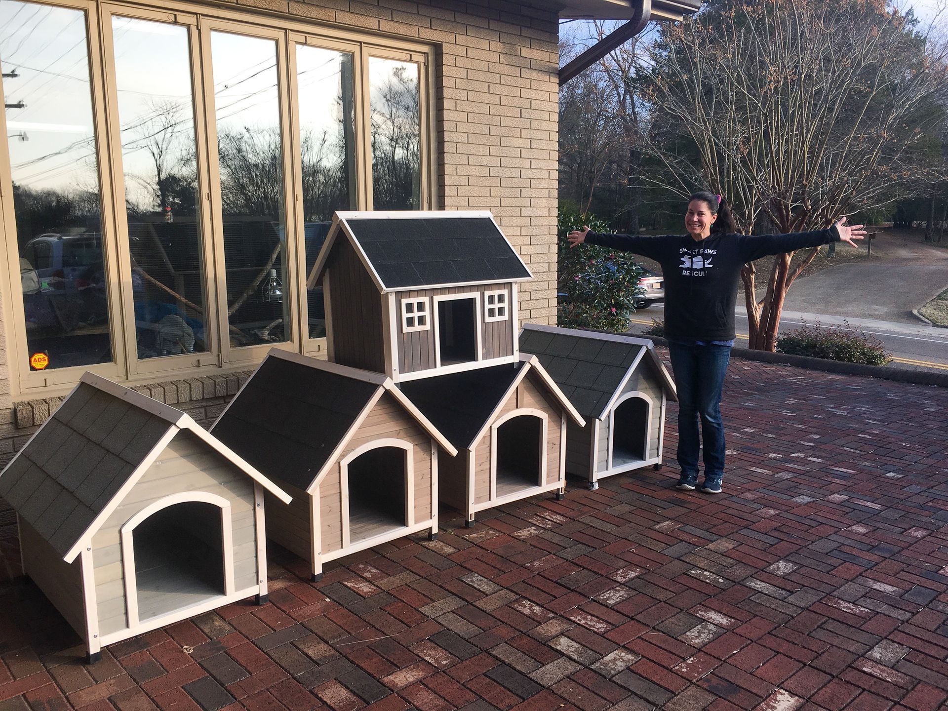 A person stands with arms spread wide next to a complex arrangement of four connected wooden dog houses on a brick patio.