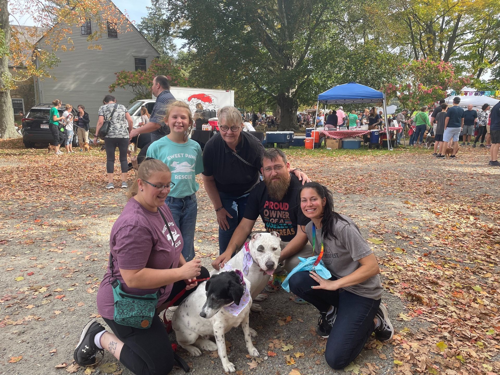A diverse group of people crouch and stand in a leafy, outdoor setting, posing with two dogs wearing purple bandanas.