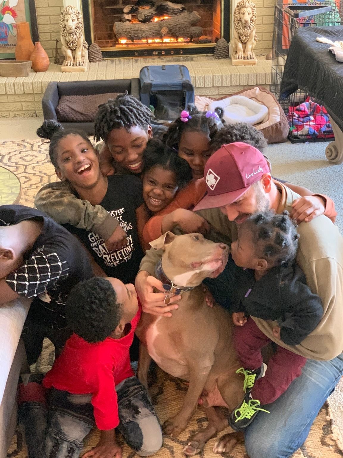 A group of children and an adult crowd around a large, light brown dog in a living room, with a lit fireplace behind them.