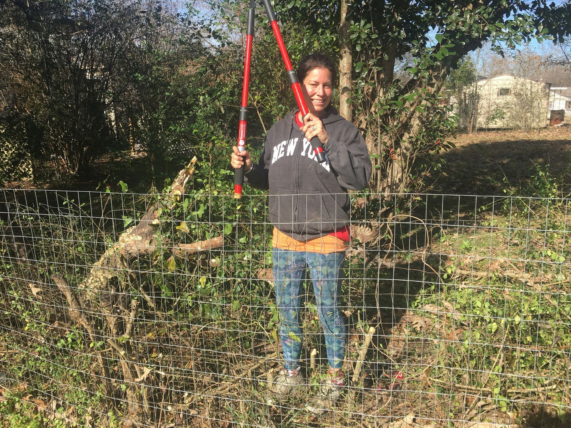 A smiling person wearing a dark hoodie and patterned leggings holds long-handled red loppers in a sunny, brushy garden.