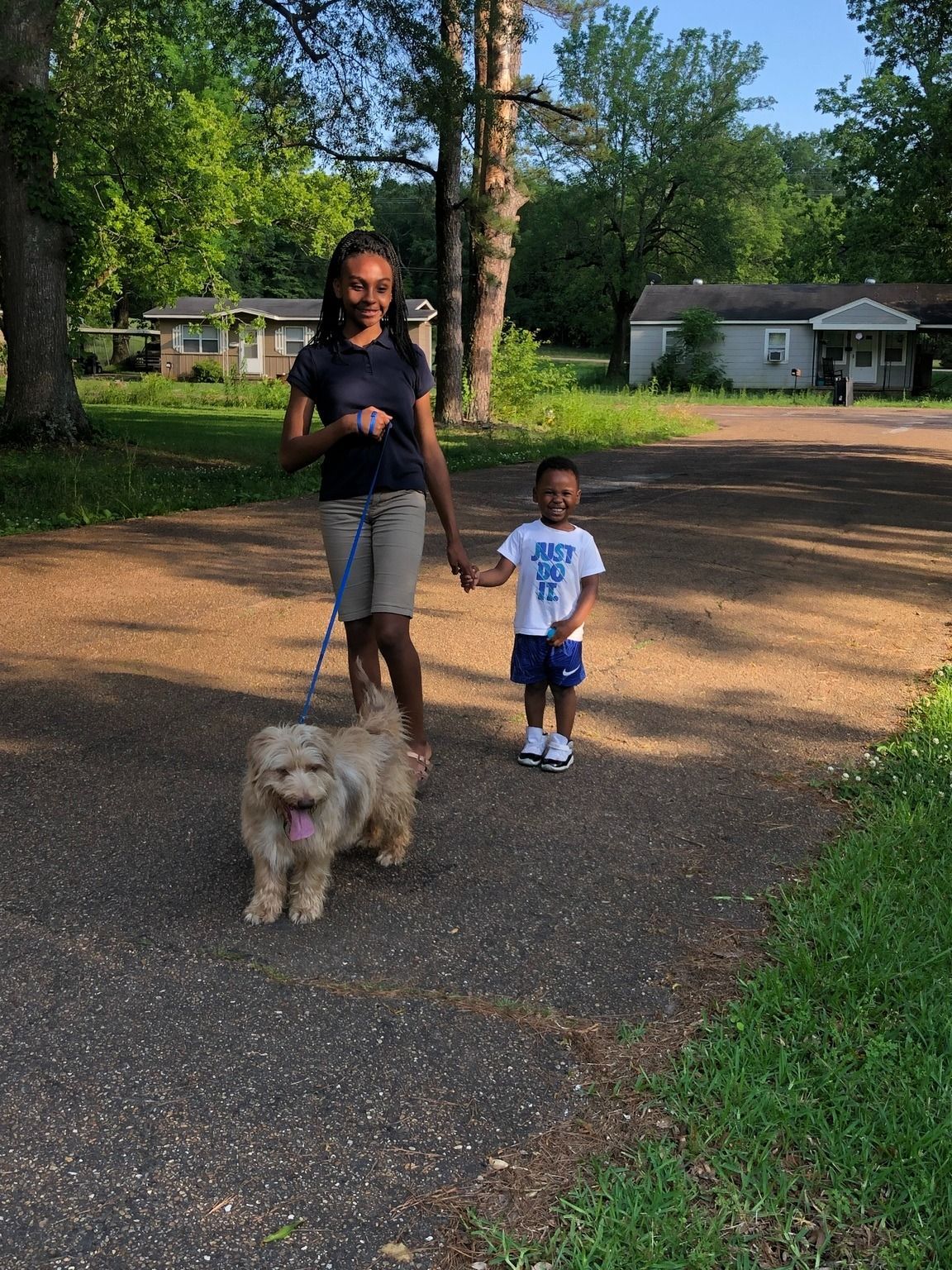 A person walks a dog and holds hands with a child on a paved path, houses and trees in the background.