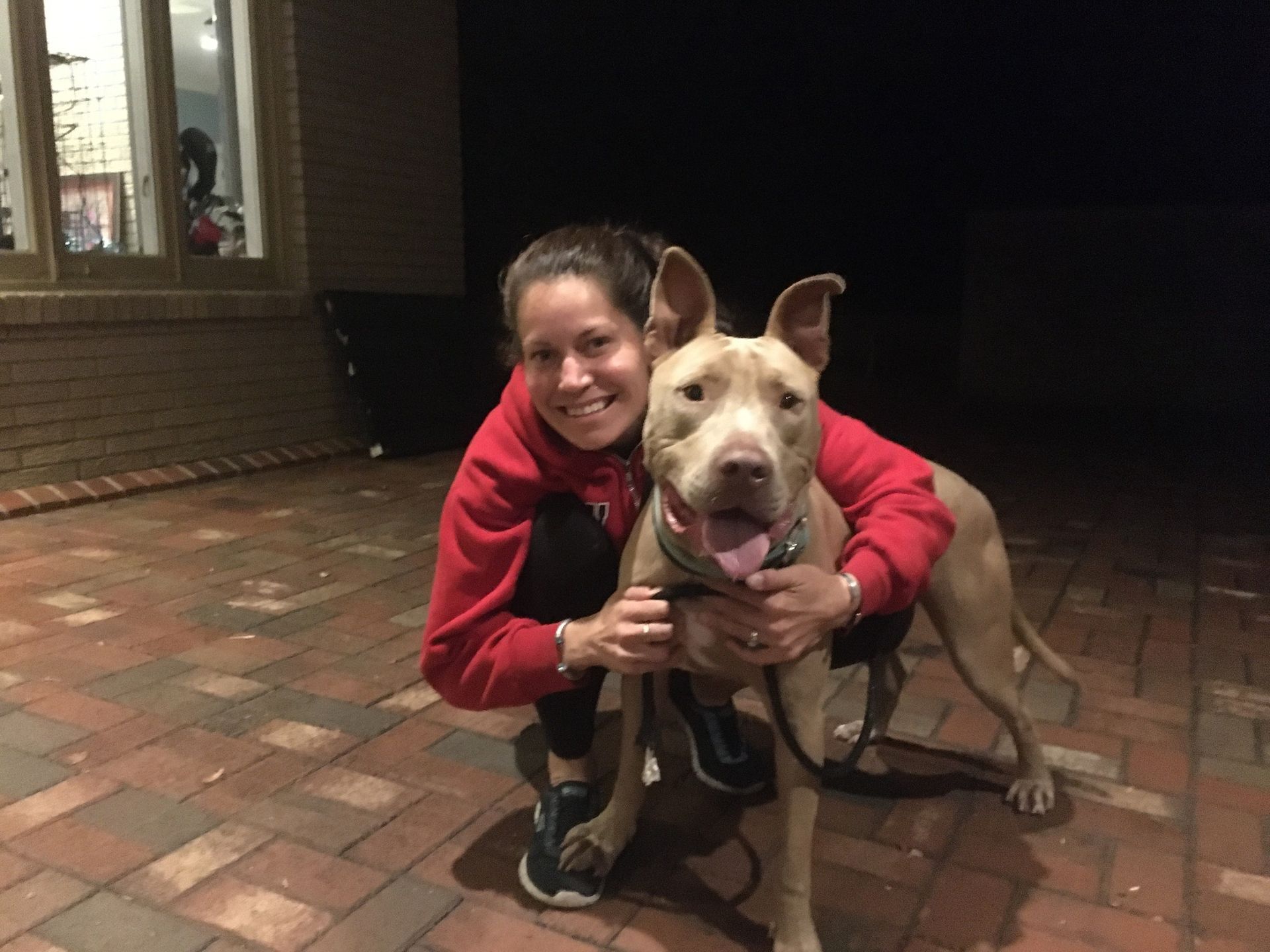 A person in a red hoodie kneels on a brick patio, smiling while hugging a tan, short-haired dog with upright ears.