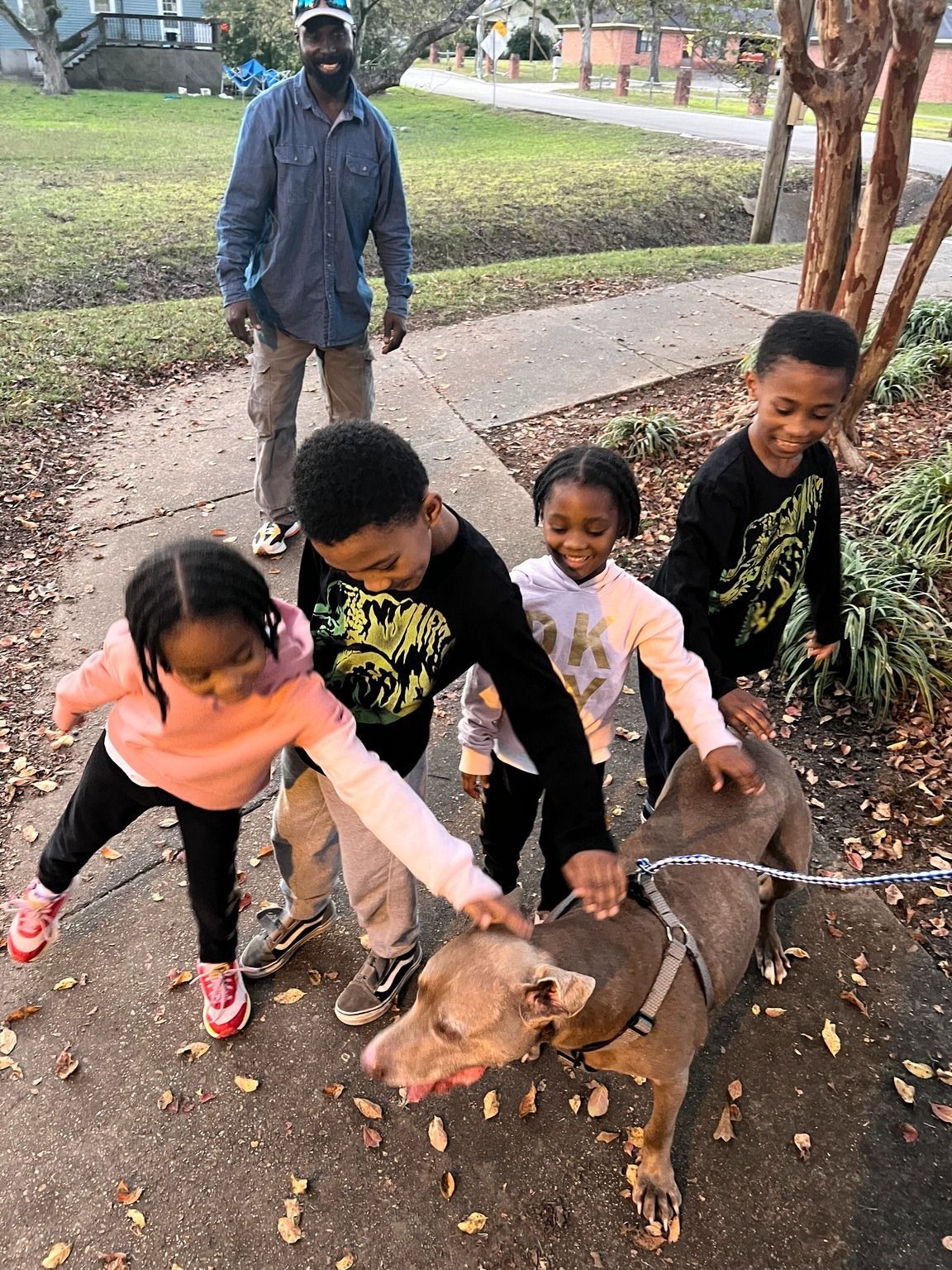 Children petting a dog on a sidewalk, an adult is nearby.