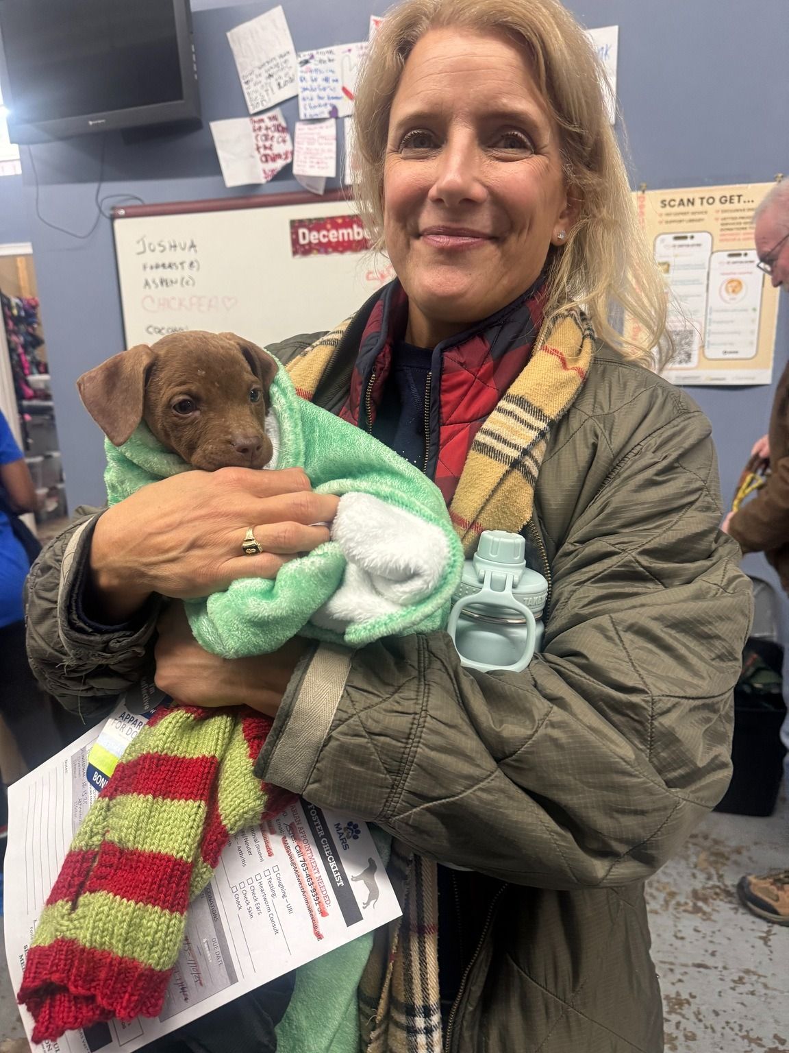 Woman holding a brown puppy wrapped in a green towel. She's inside a building and smiling.