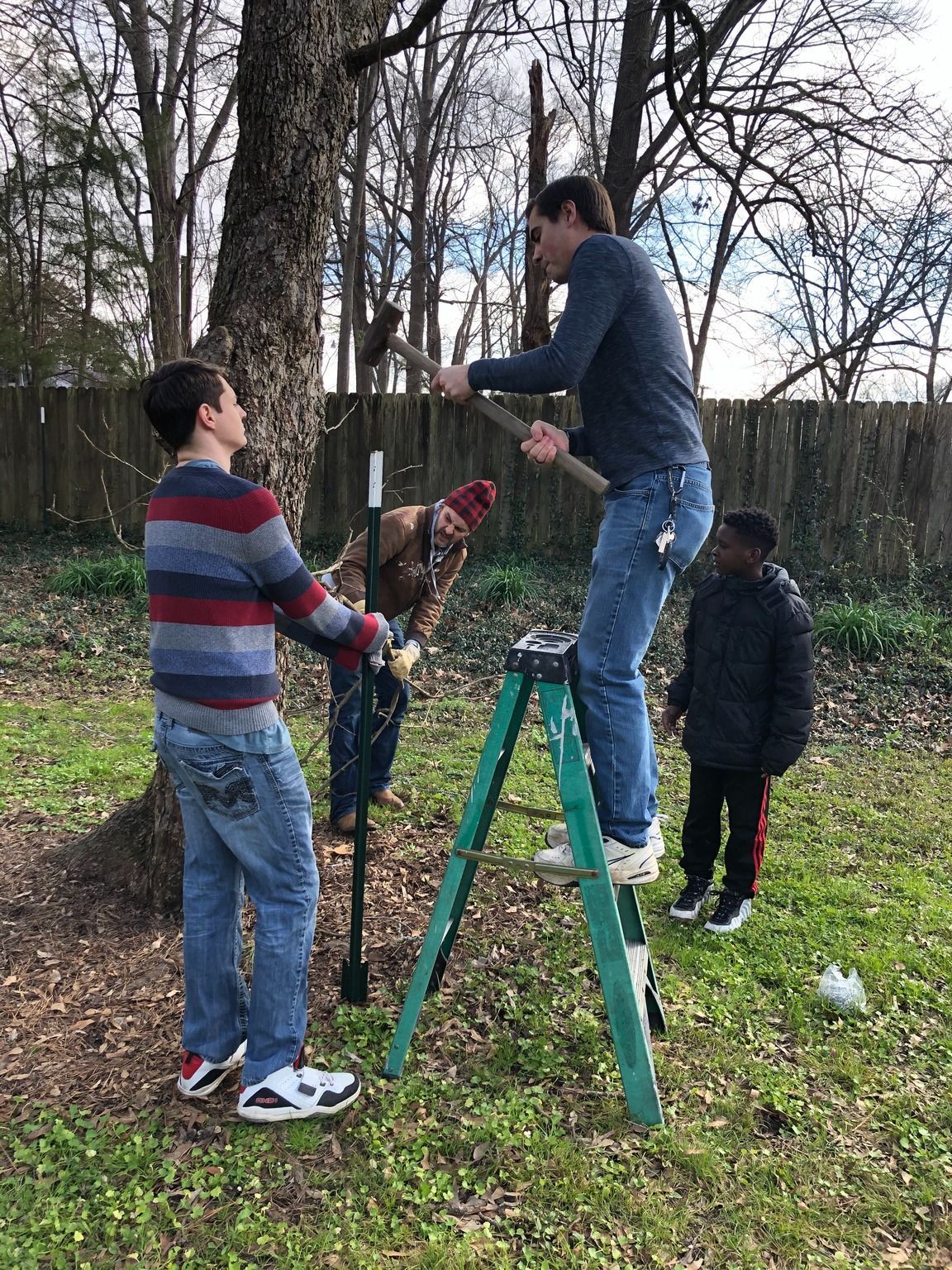 Four people working outdoors: one on a stepladder hammers a post into the ground near a tree, while three others assist.