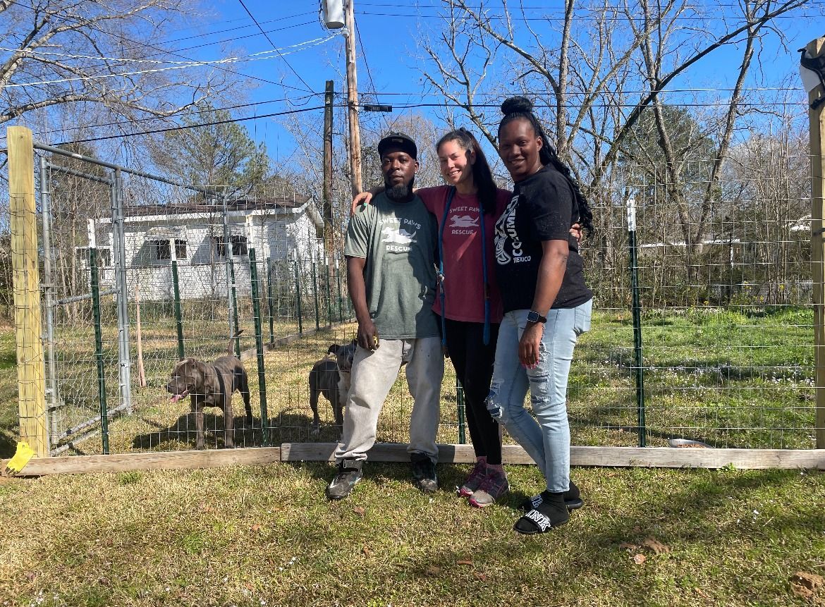 Three people stand arm-in-arm in front of a wire-fenced outdoor garden with two dogs nearby on a sunny day.